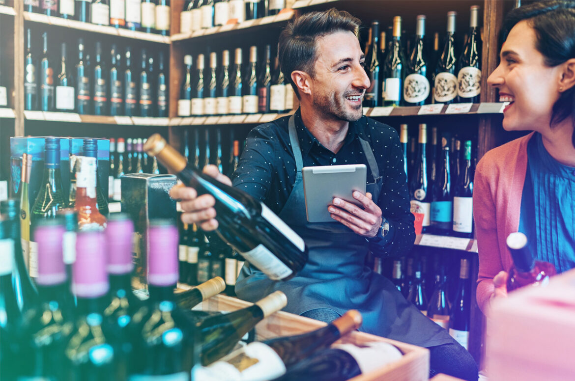 A liquor store business owner is holding a wine bottle while discussing with a client.