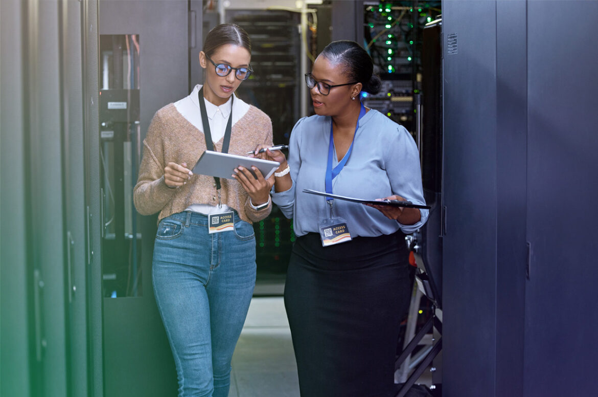 A managed service provider business owner is holding a notepad while discussing with a client in an IT closet.