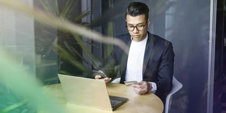 A male sitting at a table with his laptop looking at his checking account debit card while reviewing his fraud protection.